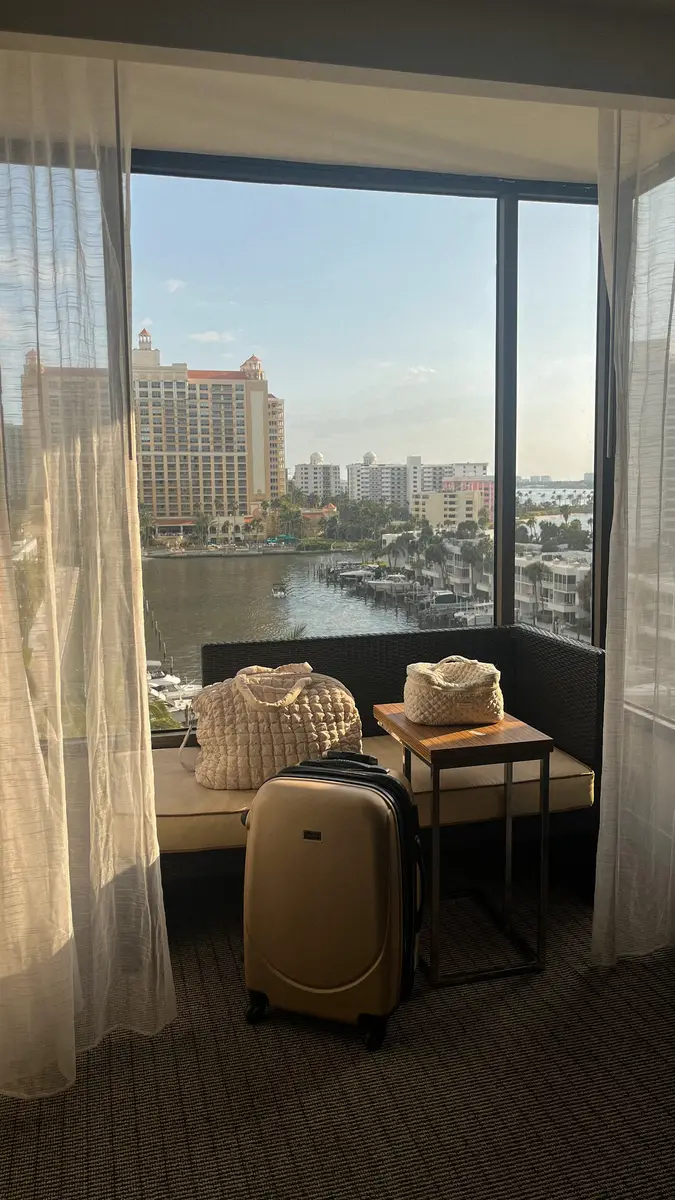 Corner of a hotel room that is al windows, with a stunning view of a marina and hotel across the marina, in the room, you can see a carry-on, a toiletry bag and a personal item bag.