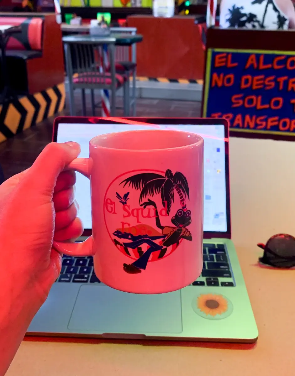 Hand holding out an El Squid Roe mug in front of a laptop while working at their venue. 