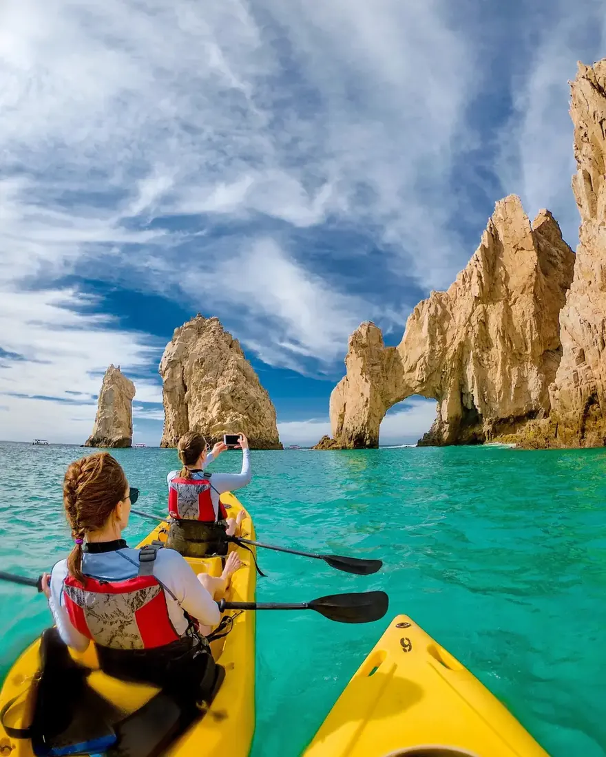 Two women riding a kayak towards the Arch of Cabo, the one in front taking a picture. 
