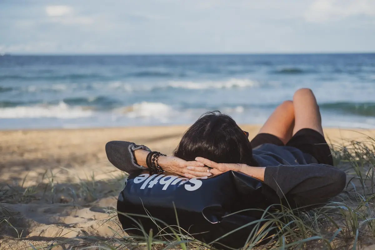 Person resting on the beach with their head on their backpack.