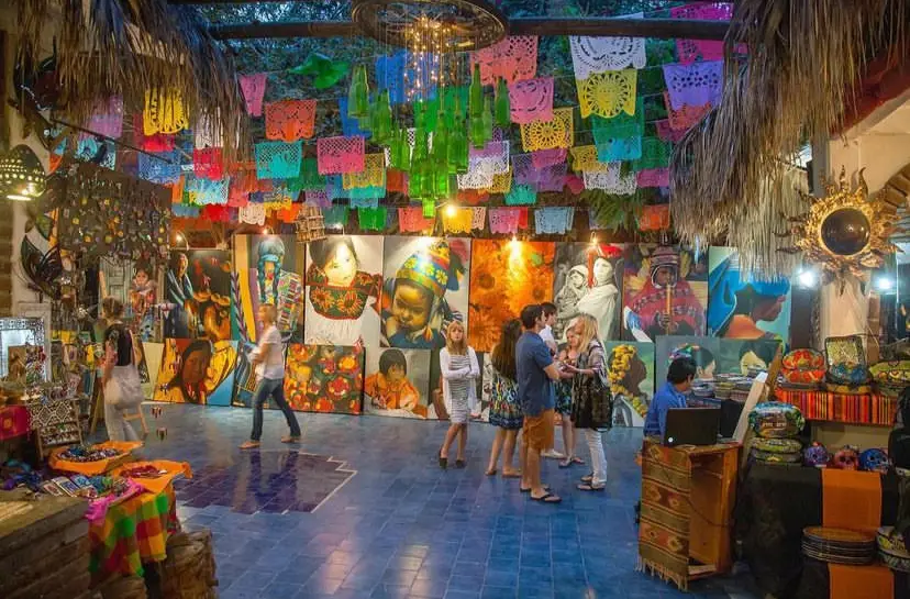 Room with walls covered in art pieces, from metalwork to paintings, with papel picado hanging from the ceiling, and various patrons walking around, looking at the art in the San Jose del Cabo Art Walk. 