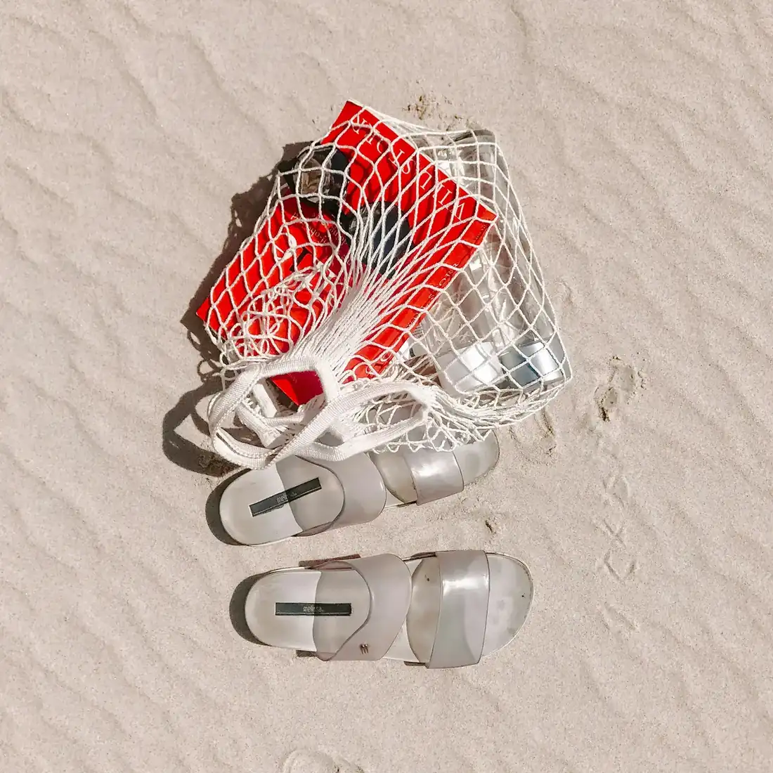 Net bag with a book and bottles of water in it and a pair of sandals next to it, resting on the sand. 