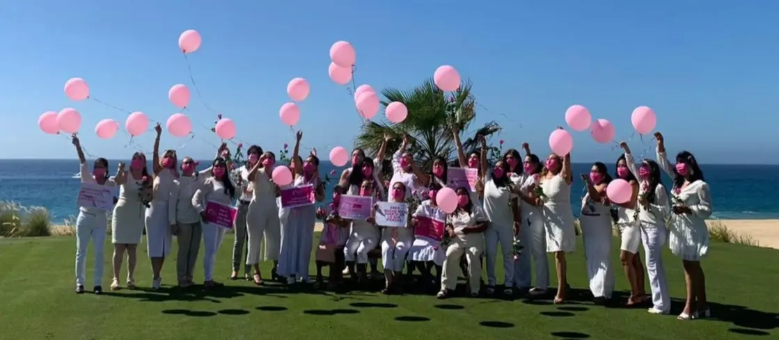 Group of Women and the Breast Cancer Society Foundation standing on a grassy clearing in front of the beach, releasing pink balloons.