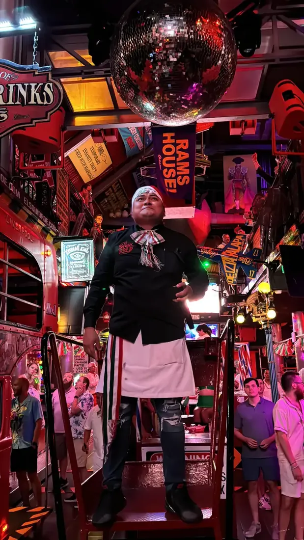 MC Iguano standing at the podium at El Squid Roe as party goers dance around him celebrating Mexico's Independence Day. 