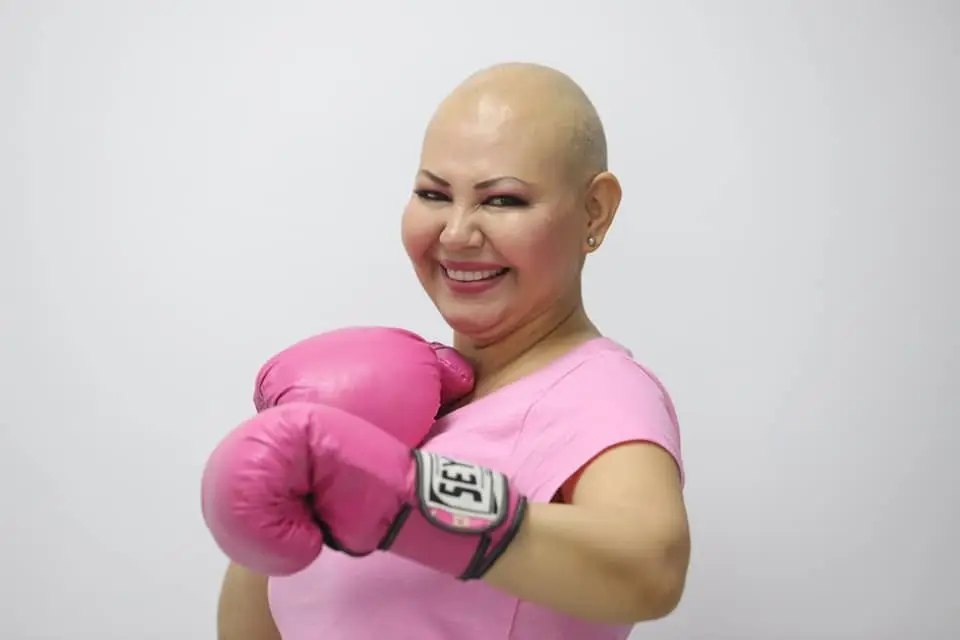 Woman with no hail, dressed in pink, with pink boxing gloves help up in fighting position.