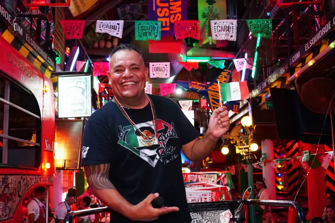 Mc Pablo dressed in red, white, and green, holding a little Mexican flag, the venue behind him decorated in the same colors. 