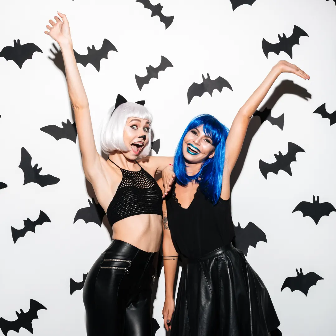 Two girls posing in front of a white wall decorated with bats, one of the girls is dressed up as a cat and the other is dressed in all black and has a blue wig. 