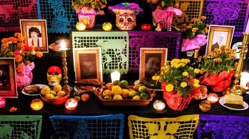 Black altar made of multiple layers covered with pictures of loved ones, bowls of food, flowers, lit candles, and papel picado decorating each level of the altar. 