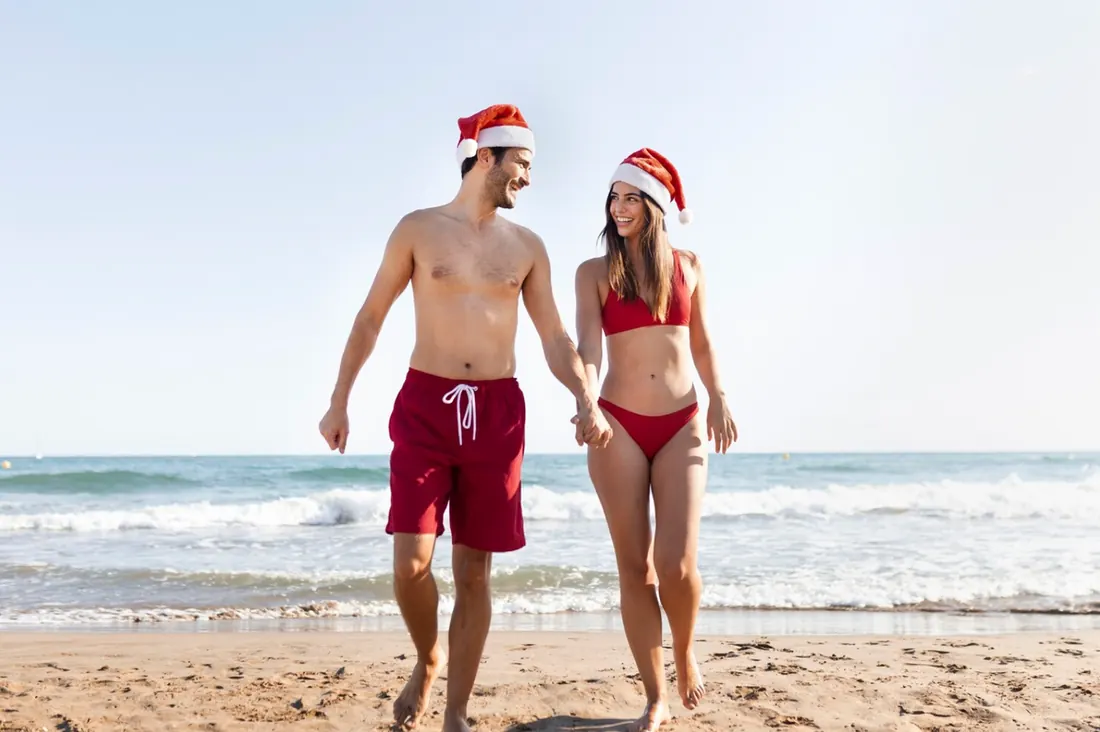 Couple walking on the beach, holding hands, wearing red swimsuits and Santa hats.