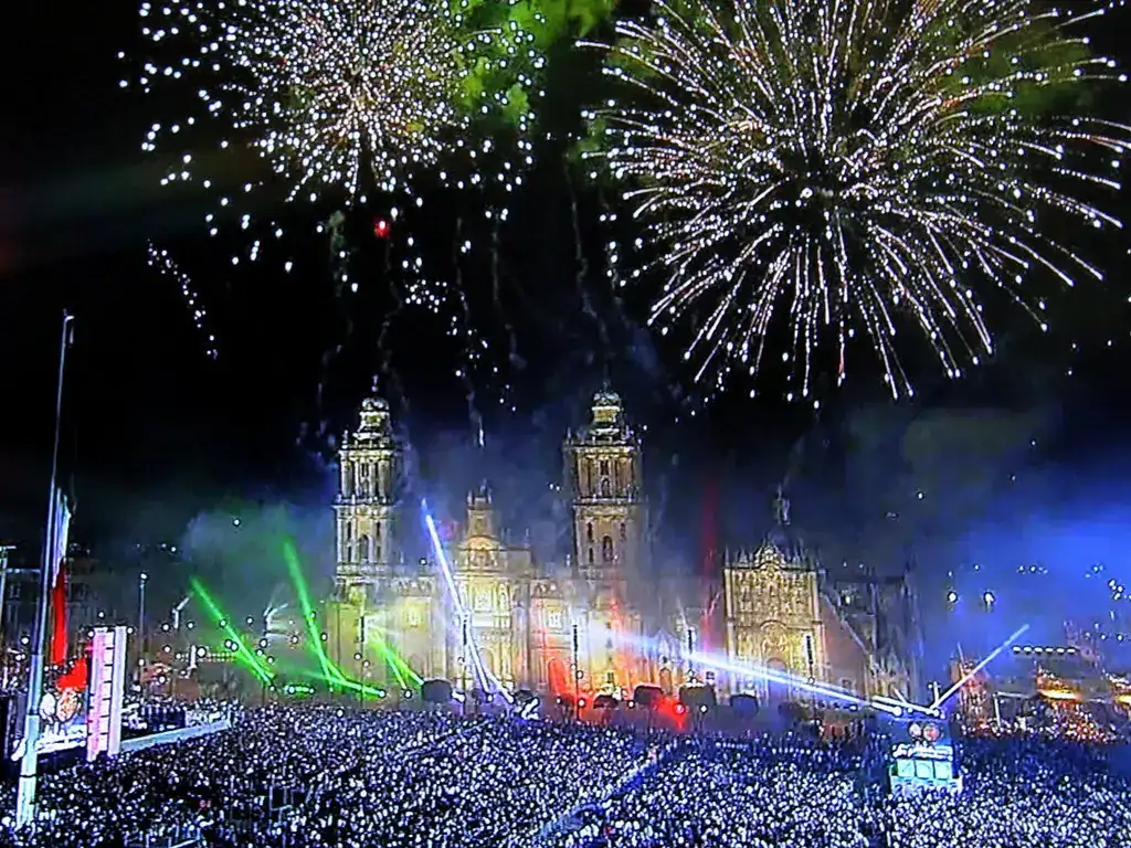 Celebration in Mexico City, with red, white, and green lights, fireworks in the sky and a massive crowd.