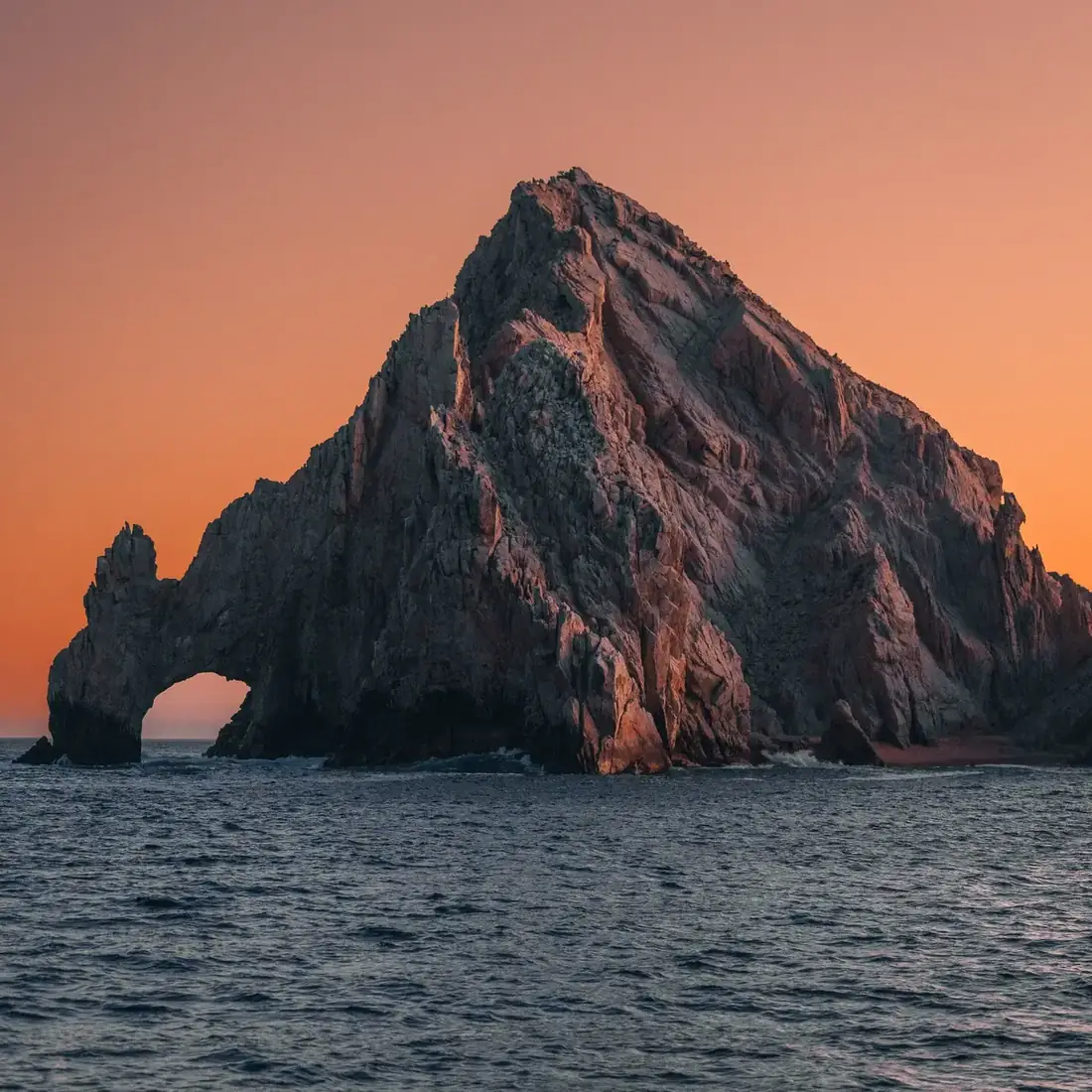 El Arco, rock formation at the tip of Baja California peninsula seen at sunset with an orange sky behind it. 