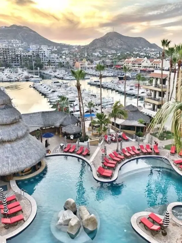 Flower shaped pool surrounded by red lounging chairs, and a palapa pool bar. Marina filled with boats and hills in the background. 