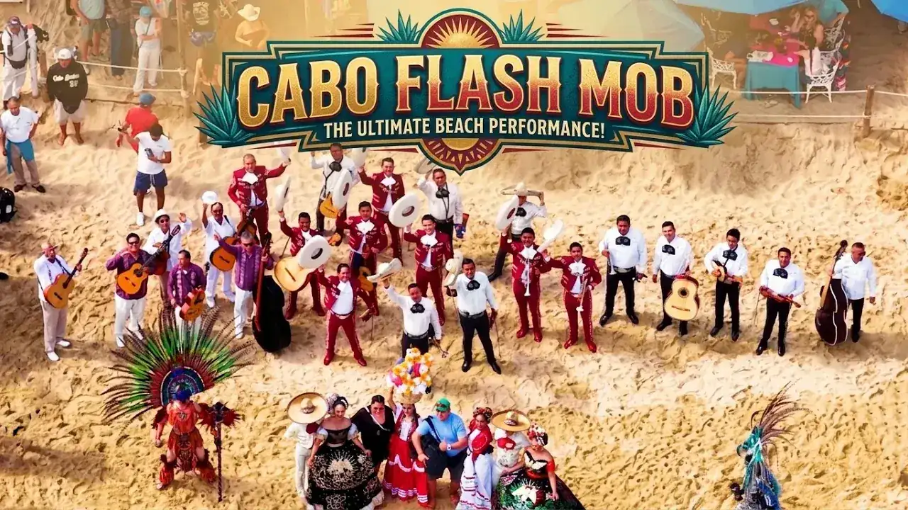 Musicians in red and white outfits perform on a sandy beach under a "Cabo Flash Mob" banner. An audience watches and colorful costumes are seen.