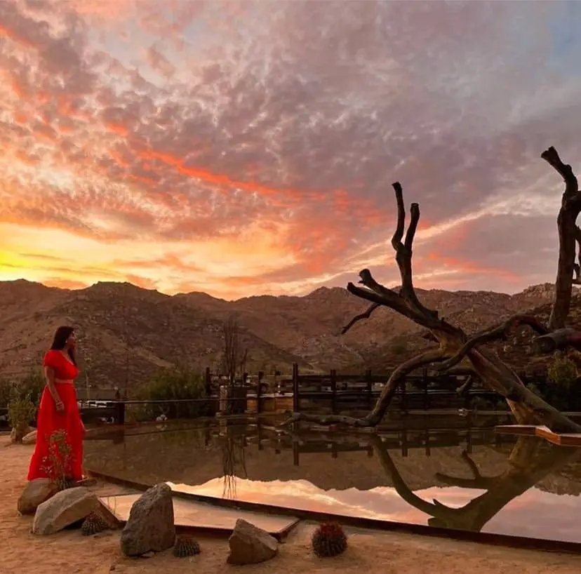 Woman looking towards a pool with the sun setting over desert hills in the background.