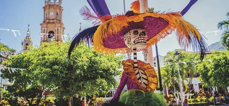 Huge statue of "La Catrina" dressed in purple with orange accents, feathers in her har, standing in front of some greenery in front of a church.