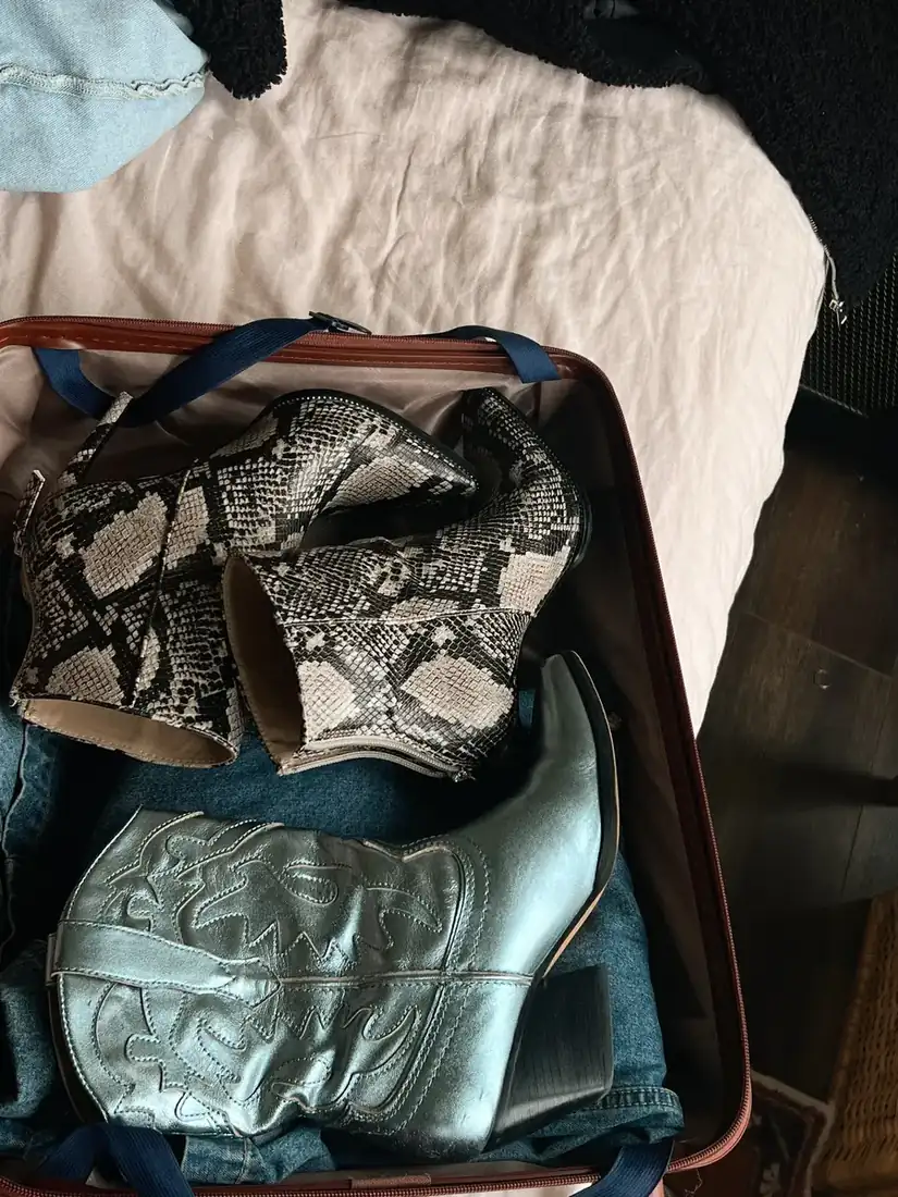 Carry on on the corner of a bed with clothes and a pair of boots in it.