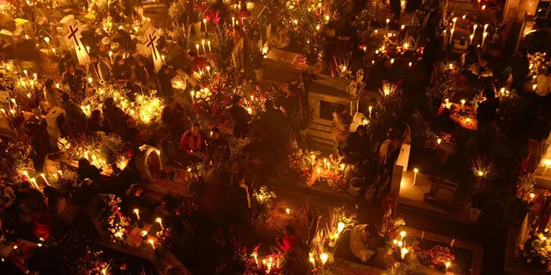 Scene of a cemetery seen from above, multiple families gathered by their loves ones tombs, illuminated by candles.