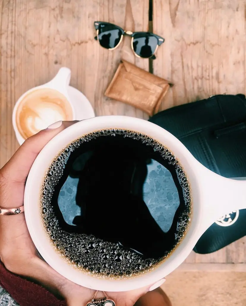 Hand holding a cup of coffee seen from above, cappuccino, wallet, purse, and sunglasses in the background.