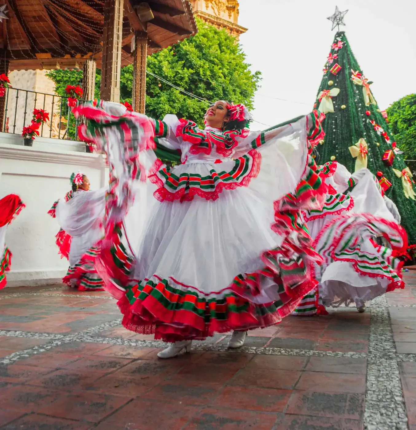 Female dancers dressed in traditional Mexican dresses with red and green accents dancing in the town square next to a large Christmas tree in Cab o, Mexico.