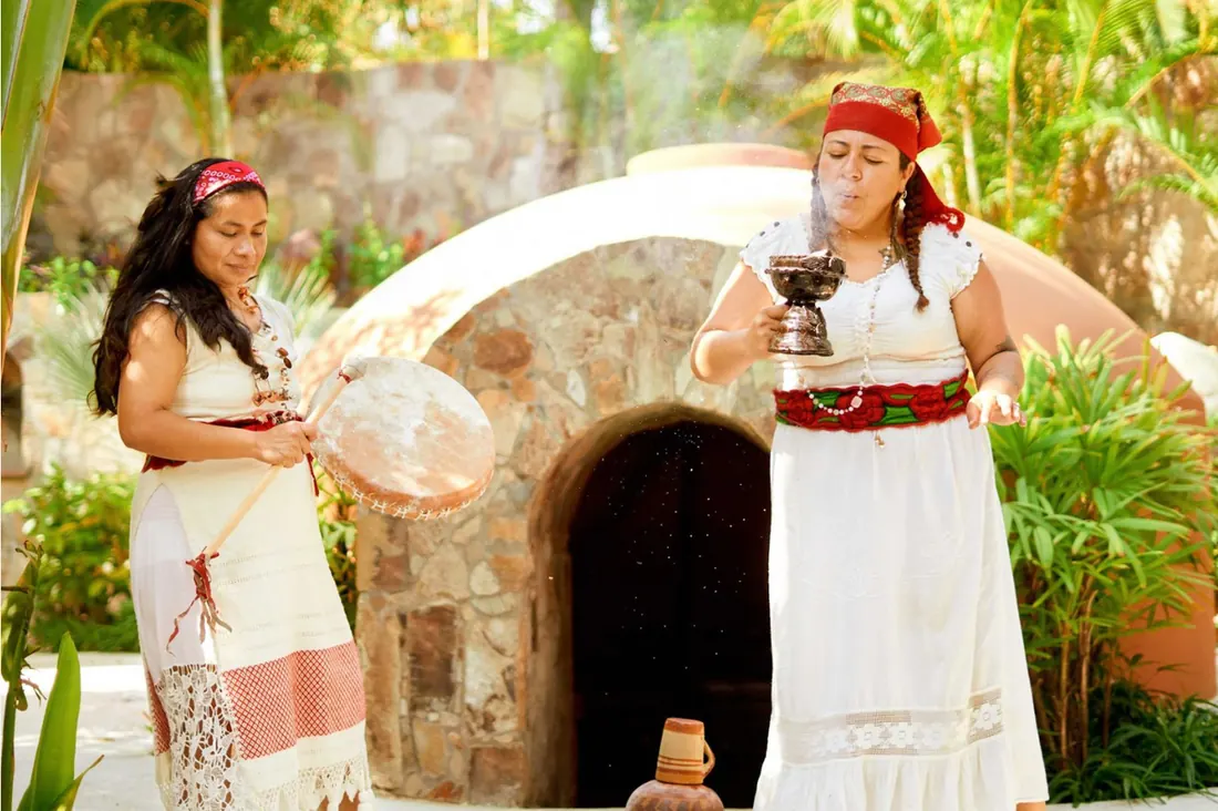 Two curandera women, one banging a drum and the other blowing on a smoking bowl, surrounded by greenery.