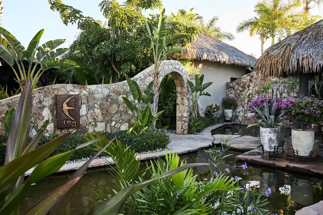 Entrance to spa by a pool and a palapa, surrounded in greenery.