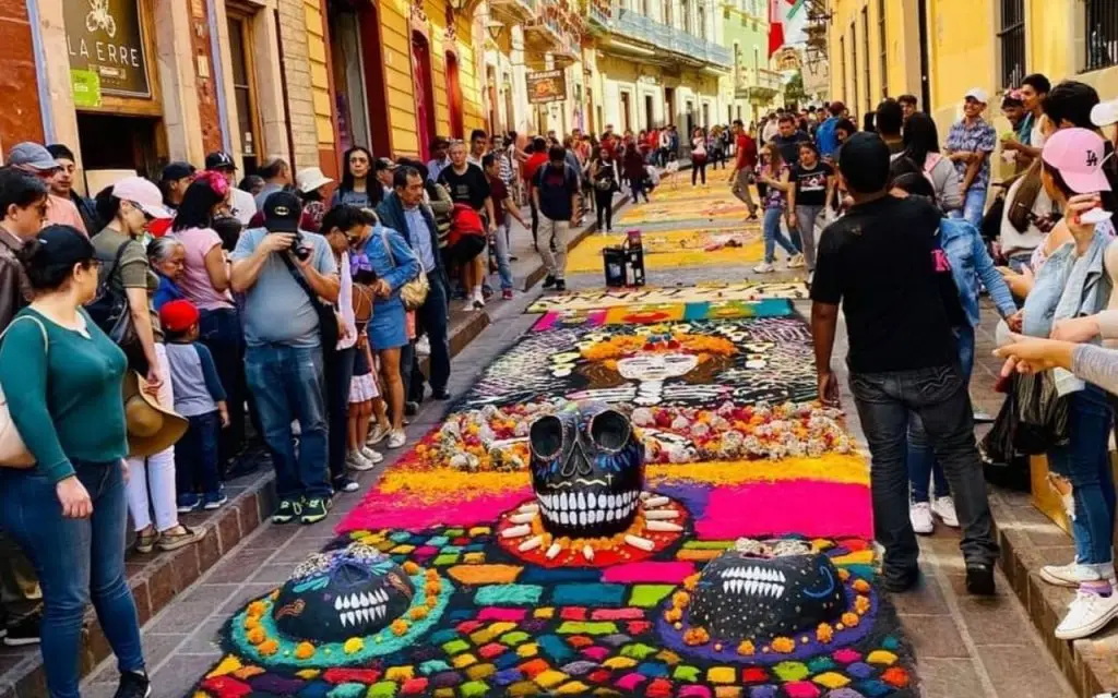 Crowd gathered on either side of the colorful "Tapete de Muerte" with colorful shapes and large skulls.