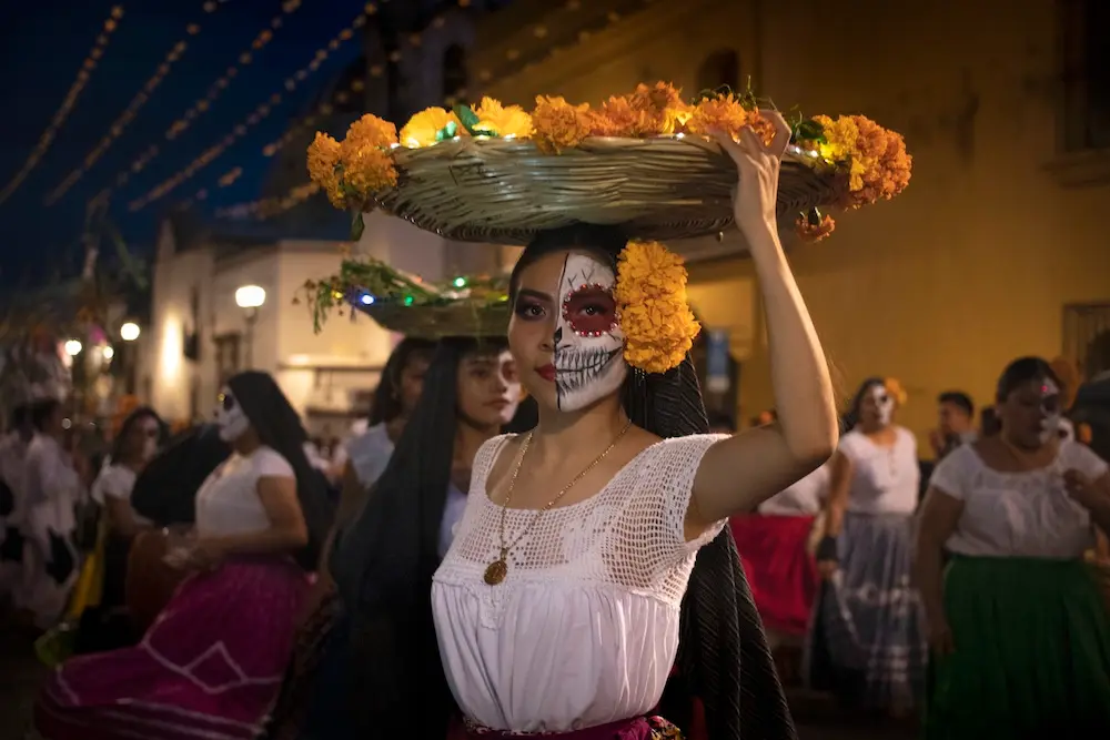 Woman with half her face painted as a skeleton, holding a basket of flowers over her head.