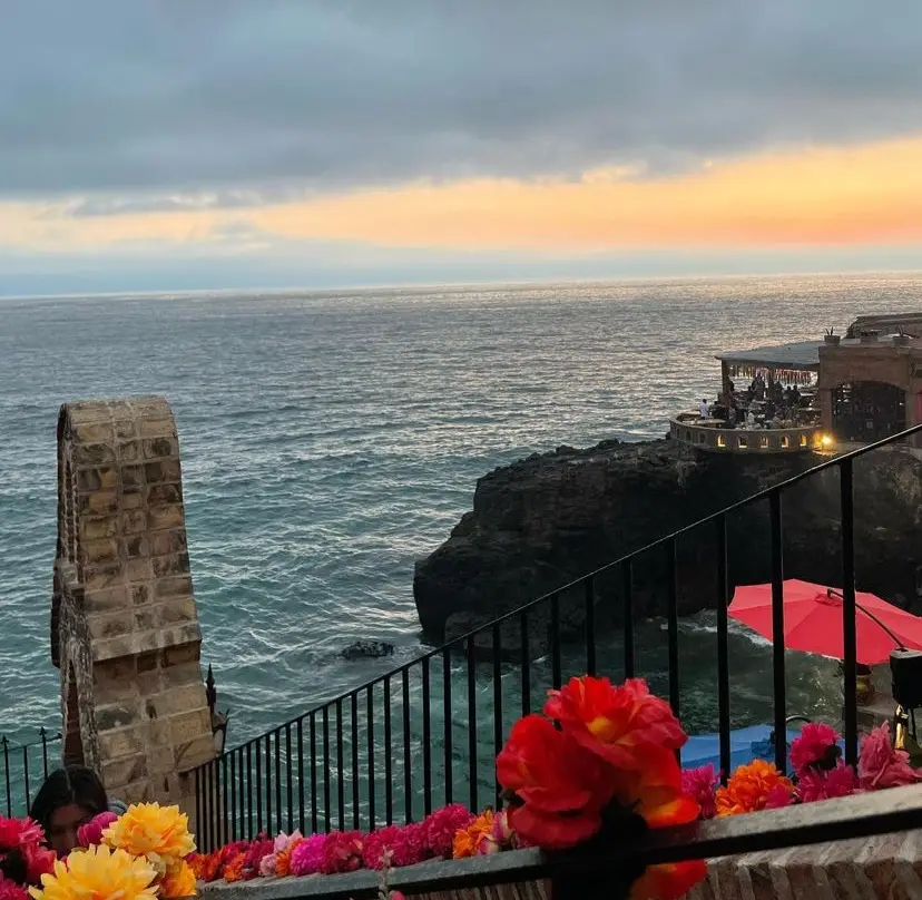 Sun setting over the ocean with a restaurant over a cliff and red flowers lining the railing of steps leading towards the water.