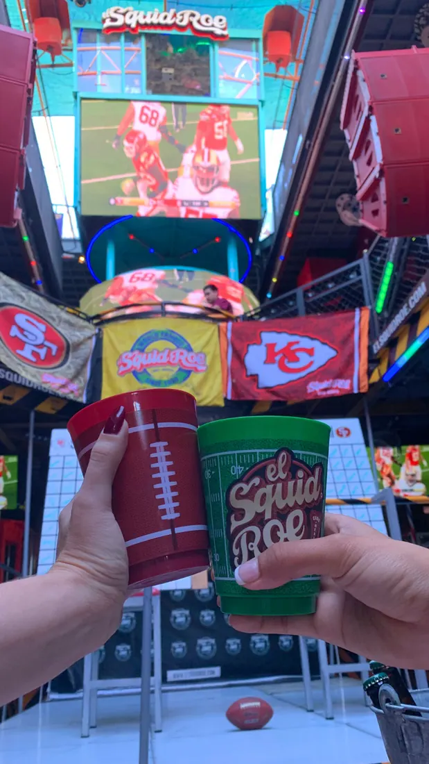 Two women holding our cups from El Squid Roe in front of the LED screesns at El Squid Roe playing the football game in Cabo, Mexico.